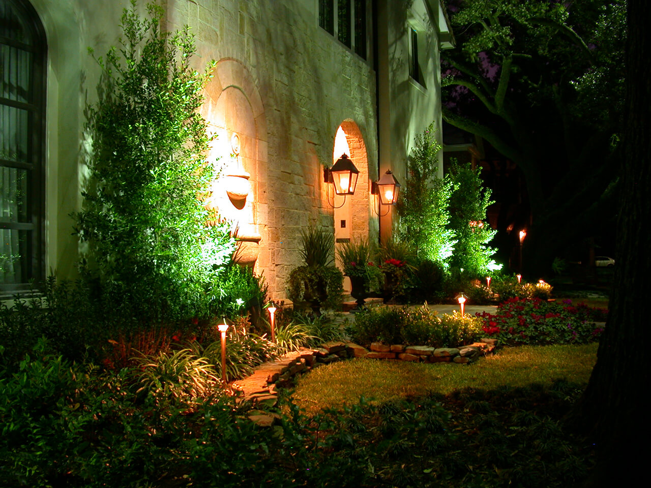 Natural Concepts light fixtures lighting the fountain, trees, and flower beds in the front of a stone house
