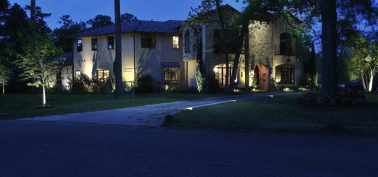 A large stone house with trees surrounding it with soft white lights going up the driveway and bright lanterns lighting the permitter of the house