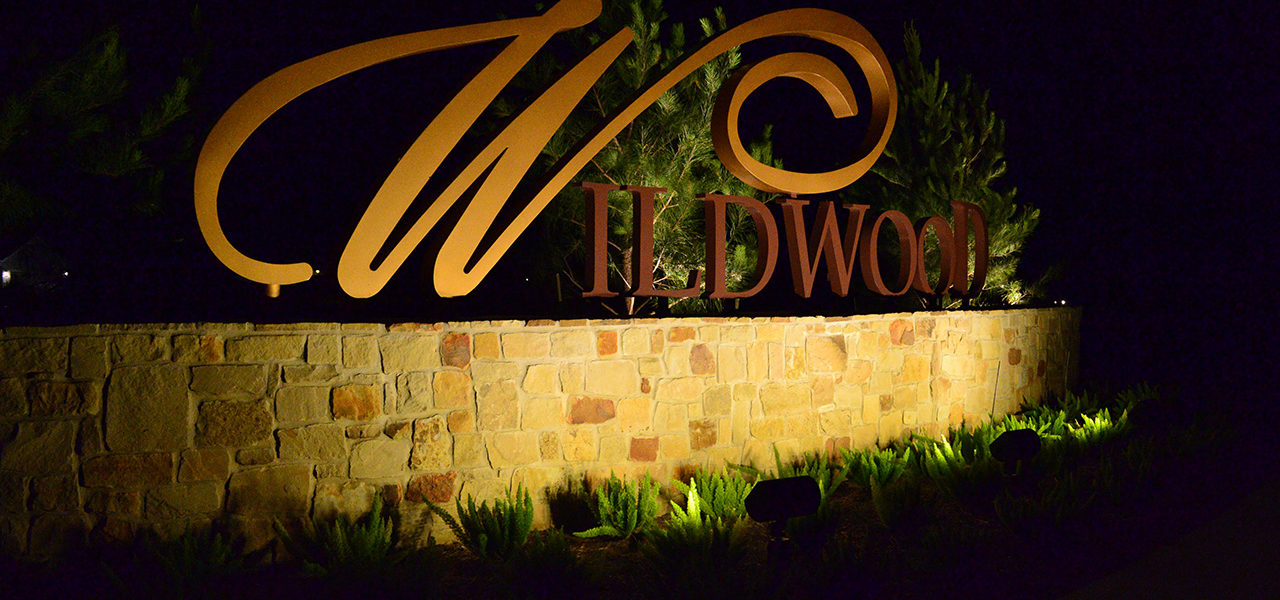 Large metal letters spelling Wildwood fitted on top of a brick wall being lighted by the lamps in the bushes below