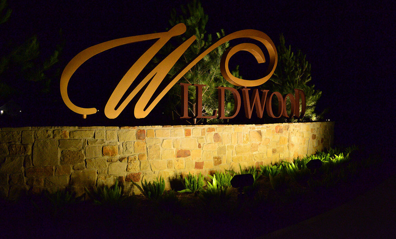 Large metal letters spelling Wildwood fitted on top of a brick wall being lighted by the lamps in the bushes below